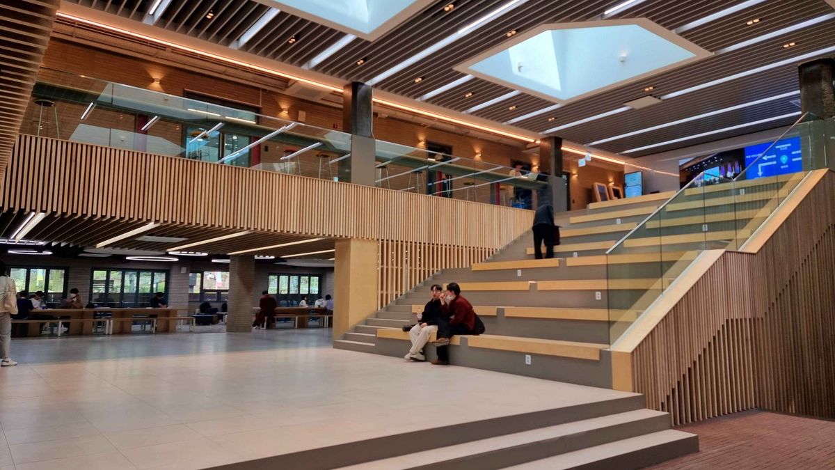 Das Bild zeigt den hellen und modernen Eingangsbereich des Unigebäudes der University of Seoul. Der Raum ist offen gestaltet mit hohen Decken, die von horizontalen, leuchtenden Elementen durchzogen sind. Eine breite Treppe mit hellen Stufen führt zu einer oberen Ebene, die durch eine Glasbrüstung begrenzt ist. Im Erdgeschoss sitzen einige Personen auf den Stufen und unterhalten sich. Im Hintergrund sind Schreibtische und Sitzgelegenheiten zu sehen, die von Studierenden genutzt werden. Große Fenster lassen Tageslicht herein und bieten einen Blick auf die Außenanlage. Der Raum wirkt einladend und lebendig.