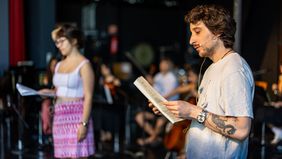 Ein junger Mann und eine junge Frau mit Textbuch in der Hand stehen auf der Bühne, im Hintergrund sind unscharf die Musiker zu sehen. 