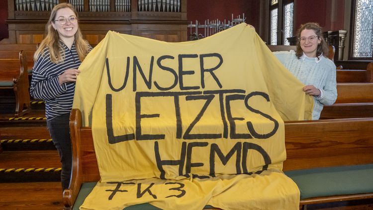 Das Bild zeigt Wencke Bammann und Mafalda Nogueira. Sie halten ein überdimensioniertes gelbes T-Shirt mit der Aufschrift "Unser letztes Hemd. Fakultät 3" hoch. Im Hintergrund sind eine Orgel und Sitzbänke des alten Lehrerseminars zu erkennen.