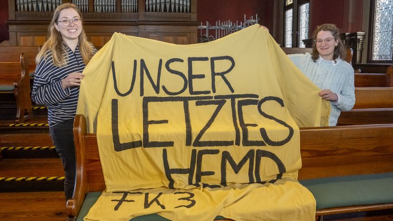 Das Bild zeigt Wencke Bammann und Mafalda Nogueira. Sie halten ein überdimensioniertes gelbes T-Shirt mit der Aufschrift "Unser letztes Hemd. Fakultät 3" hoch. Im Hintergrund sind eine Orgel und Sitzbänke des alten Lehrerseminars zu erkennen.