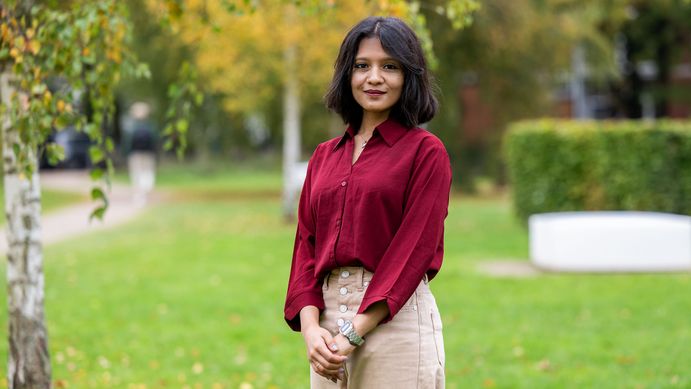 Das Bild zeigt eine junge, erwachsene, Frau, mit beiger Hose und roter Bluse. Sie steht auf einer Wiese, neben ihr ist der Stamm einer Birke zu erkennen. Sie lächelt leicht in die Kamera. Ihr Haar ist schulterlang und dunkel.