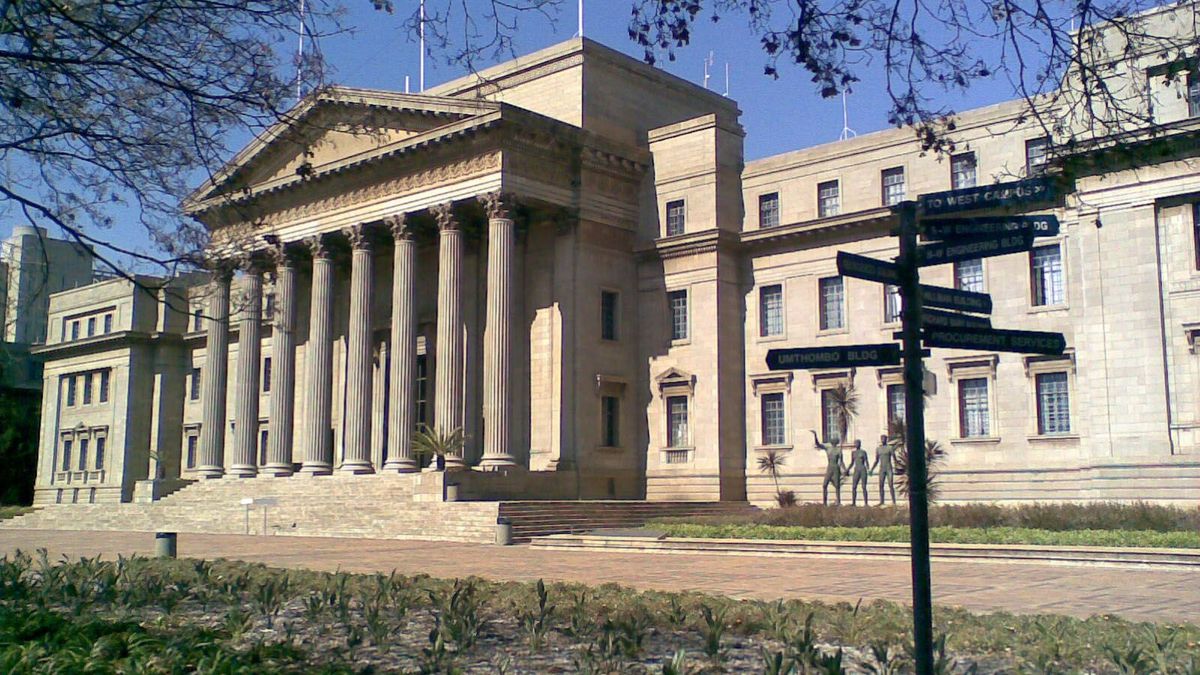 Das Bild zeigt das historische Hauptgebäude der University of the Witwatersrand in Johannesburg, Südafrika. Es ist ein imposanter, mehrstöckiger Bau im neoklassizistischen Stil mit einer Fassade aus hellen Steinquadern und einer Säulenreihe. Im Vordergrund befindet sich ein gepflegter Rasen und Wegweiser, die auf verschiedene Gebäude und Einrichtungen des Campus hinweisen.
