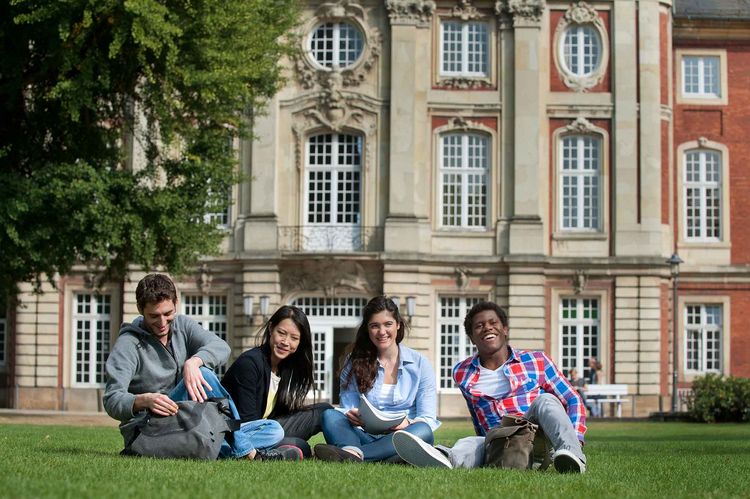 Infos für internationale Studierende Vier junge Menschen sitzen lächelnd vor einem historischen Gebäude aus rotem Backstein.