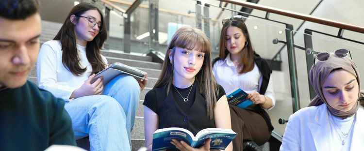 Das Bild zeigt fünf junge Studierende, die auf den Stufen einer Treppe sitzen und lernen. Vier der Personen sind Frauen, eine davon trägt ein Kopftuch und eine Person ist ein Mann. Alle lesen Bücher oder Notizen und scheinen sich in einer Lernumgebung zu befinden, möglicherweise in einer Bibliothek oder Universität.