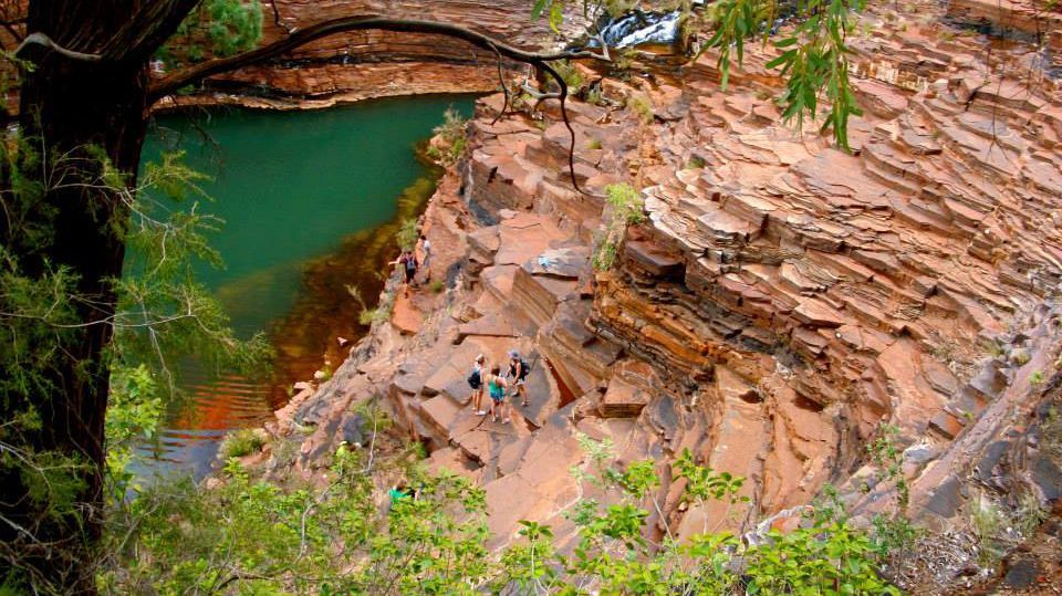 Das Bild zeigt eine Gruppe von Menschen, die an einem felsigen Abhang neben einem türkisfarbenen Wasserloch entlangwandern. Die Felsen sind rötlich-braun und weisen eine interessante Schichtung auf. Üppige Vegetation umrahmt die Szene. Die Menschen wirken klein im Vergleich zu den beeindruckenden Felsformationen. Die Szene vermittelt ein Gefühl von Abenteuer und Naturverbundenheit.
