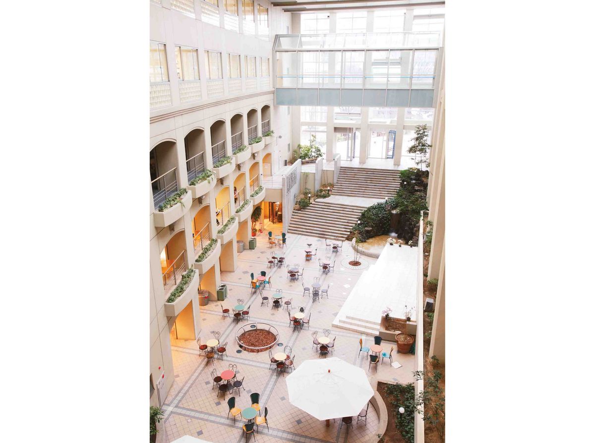 Die Galleria der Chukyo Universität auf dem Nagoya Campus, ein Innenhof eines Gebäudes mit mehreren Stockwerken, die durch Arkaden verbunden sind. Der Innenhof ist mit Tischen und Stühlen möbliert und enthält Pflanzen und Bäume. 