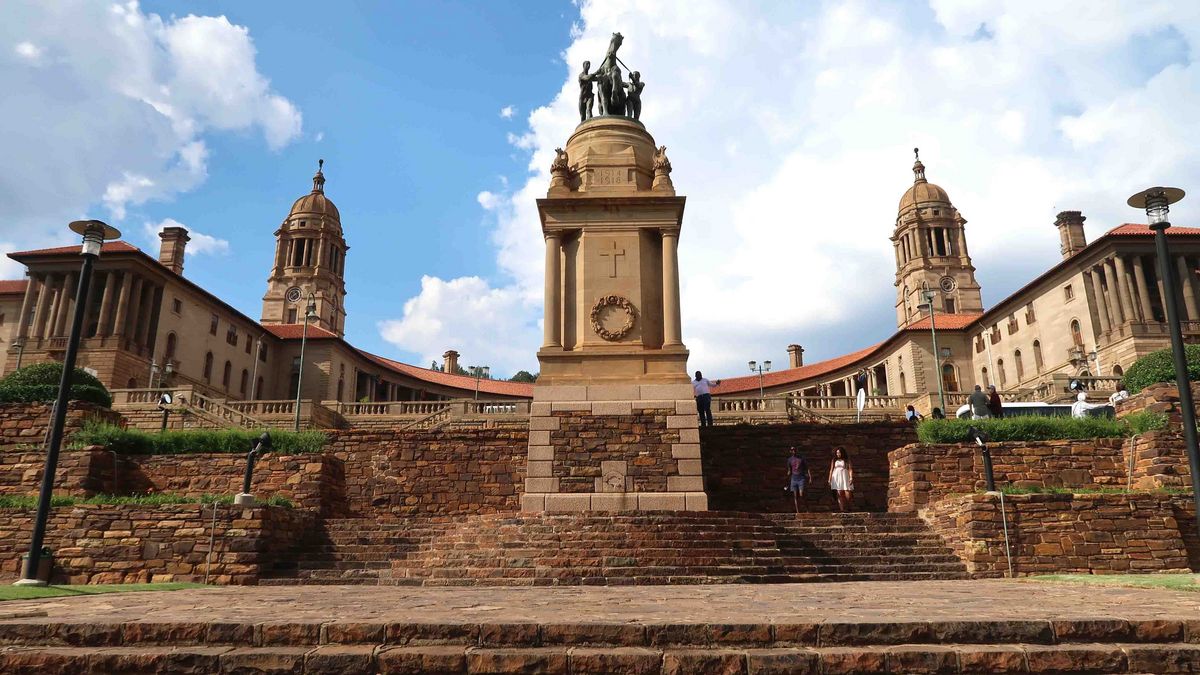 Das Bild zeigt die Union Buildings in Pretoria, Südafrika, das offizielle Regierungssitz des Landes. Das Gebäudeensemble ist durch eine zentrale Gedenkstätte mit einer Statue und zwei markante Türme gekennzeichnet. Die Union Buildings stehen auf einem Hügel und sind von gepflegten Grünflächen und Treppen umgeben. Mehrere Personen sind auf dem Gelände zu sehen, die das Gebäude erkunden oder passieren. Der Himmel ist blau mit vereinzelten Wolken.