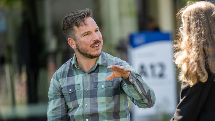 Sebastian Wilks is wearing a blue and green checked shirt and is sitting on a bench in front of building A12. He has short dark hair and a beard. He is gesturing with his left hand. Opposite him is a young woman who is interviewing him. The woman is only visible from behind; she has shoulder-length, blonde, curly hair.