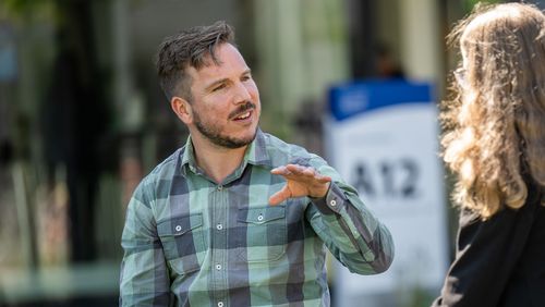 Sebastian Wilks is wearing a blue and green checked shirt and is sitting on a bench in front of building A12. He has short dark hair and a beard. He is gesturing with his left hand. Opposite him is a young woman who is interviewing him. The woman is only visible from behind; she has shoulder-length, blonde, curly hair.