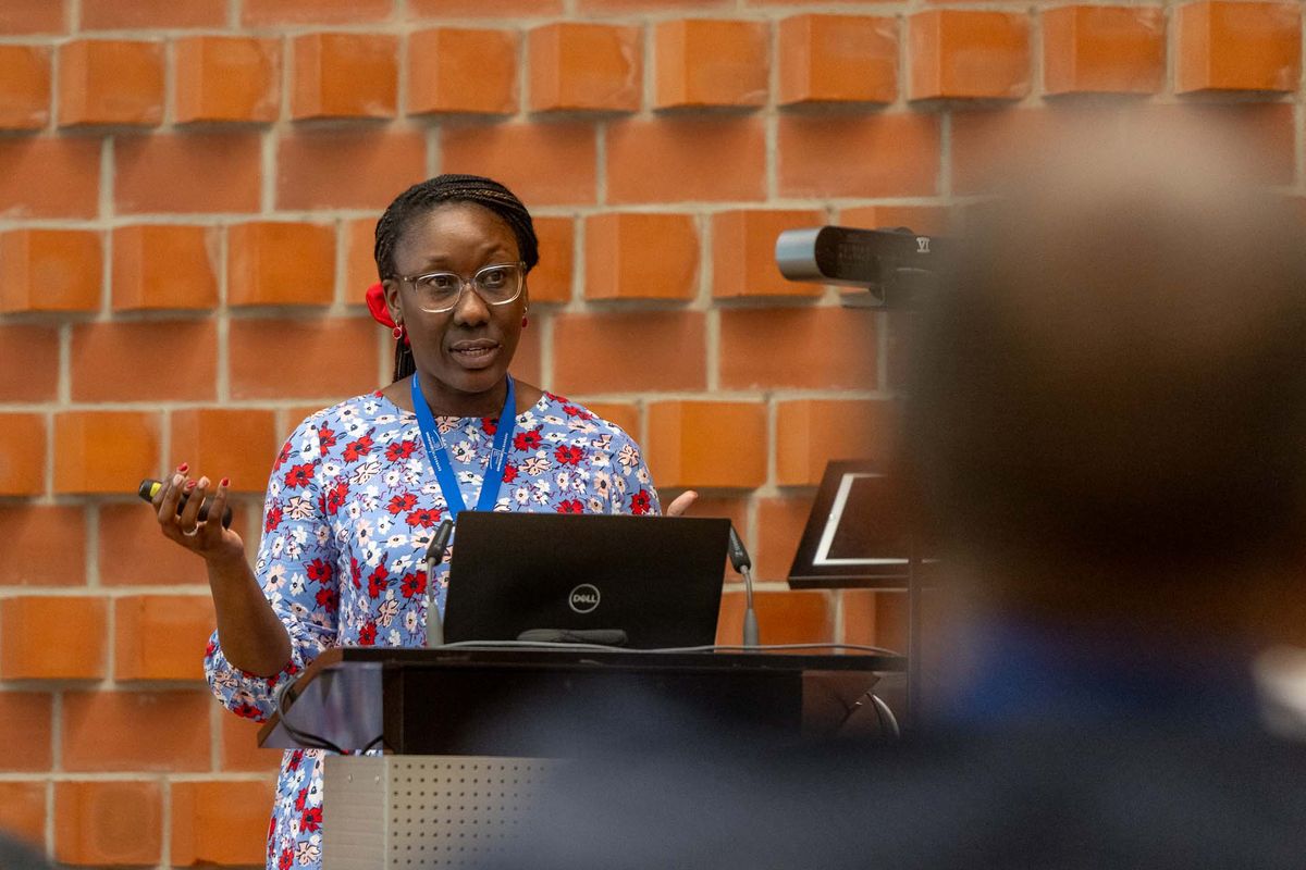 Symposium Eine Frau mit geflochtenem Haar und Brille steht hinter einem Pult und hält einen Vortrag. Sie trägt ein geblümtes Hemd und eine Lanyard um den Hals. Auf dem Pult steht ein Laptop. Im Hintergrund ist eine rote Backsteinwand und ein Mikrofon zu sehen. Das Bild vermittelt den Eindruck einer Präsentation oder eines Vortrags während des Symposiums. Die Frau scheint selbstbewusst und engagiert zu sein.