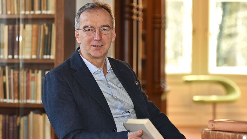 Das Bild zeigt Matthias Bormuth. Er sitzt mit einem Buch in der Hand vor einem Bücherregal im Karl-Jaspers-Haus und blickt lächelnd in die Kamera.