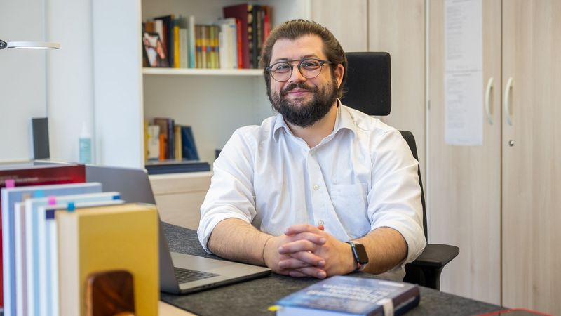 Das Bild zeigt Alexander Max Bauer. Er sitzt an einem Schreibtisch, hinter ihm steht ein Bücherregal. Vor ihm liegt ein dickes Buch, zudem ist sein Laptop aufgeklappt. Er schaut in die Kamera.