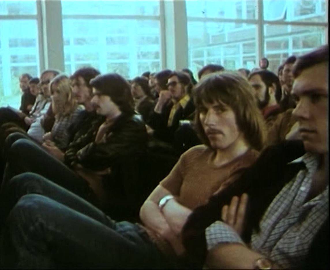Das Bild ist ein Screenshot aus einem Imagefilm der Uni. Es zeigt Studierende, die in der Aula in A 11 sitzen und mit verschränkten Armen einem Vortrag lauschen. Einer der Männer schaut leicht genervt in die Kamera.