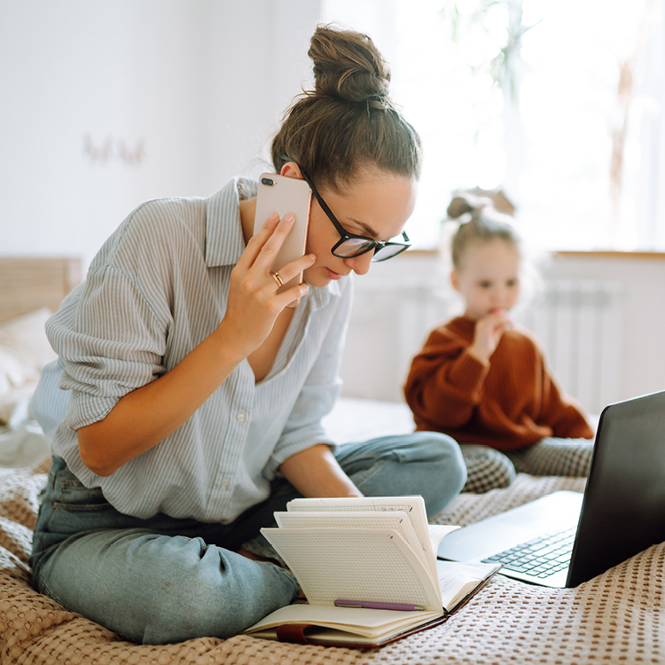 Vereinbar mit Familie, Job und Freizeit Frau arbeitet mit Laptop und Notizbuch im Homeoffice. Im Hintergrund ist ein Kleinkind zu erkennen.