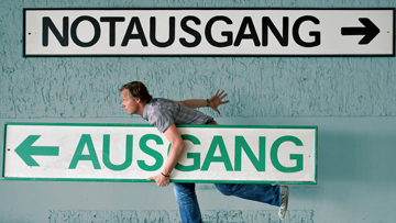 Eine Person hält ein Schild, auf dem Ausgang steht, und läuft nach links. Darüber steht ein Schild mit der Aufschrift Notausgang, das einen Pfeil nach rechts zeigt.
