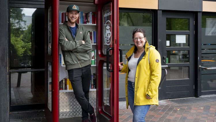 On the right of the photo is a smiling woman in a yellow jacket holding open the door to a telephone booth. In the booth, a smiling young man is standing in front of a shelf of books.